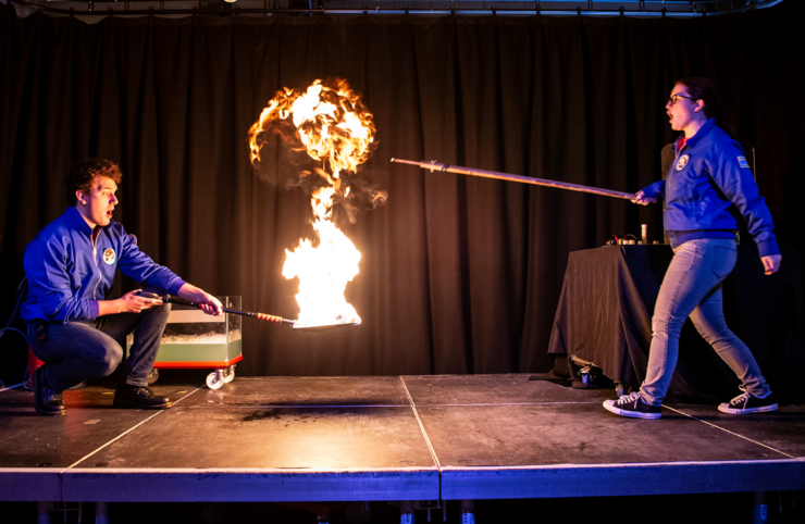 Two science Inspirers are on the science theatre stage. One of them is holding a long stick (right), the other is crouched down hold a shovel that's on fire.