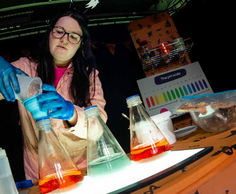 Aly, Public Curiosity Officer at WSC is wearing a white lab coat and blue rubber glovers. She is pouring chemicals into beakers.