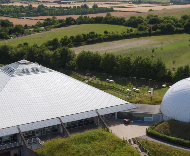 Ariel shot of Winchester Science Centre. The white roof of the iconic pyramid and dome is surrounded by green trees and fields.