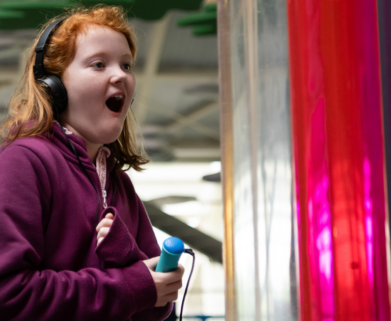 Girl with mid-length ginger hair is playing on  the Velocity exhibit. She is holding the Discovery Pen in her right hand and wearing a pair of black headphones on her head. She has an excited expression.