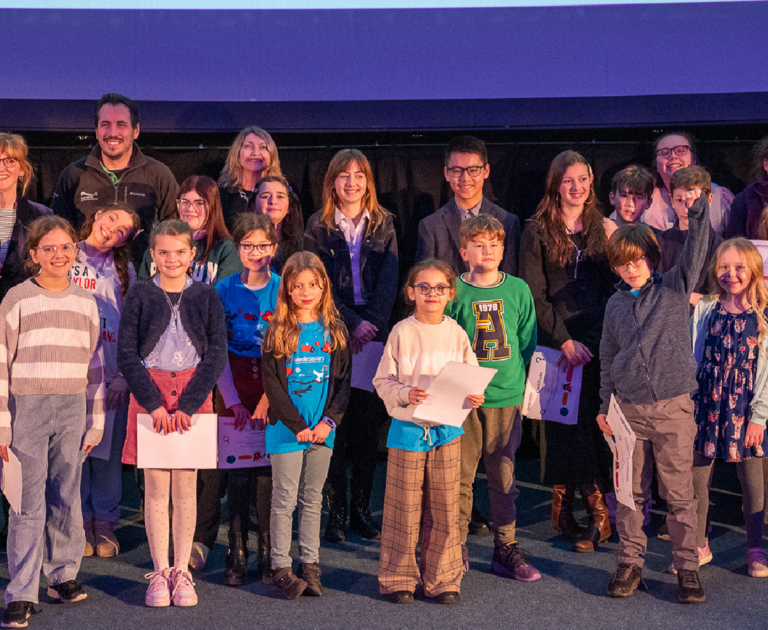 A group pf people are stood on a stage in front of a Planetarium screen. Adults are at the back and children are at the front. Some of the children are holding certificates.