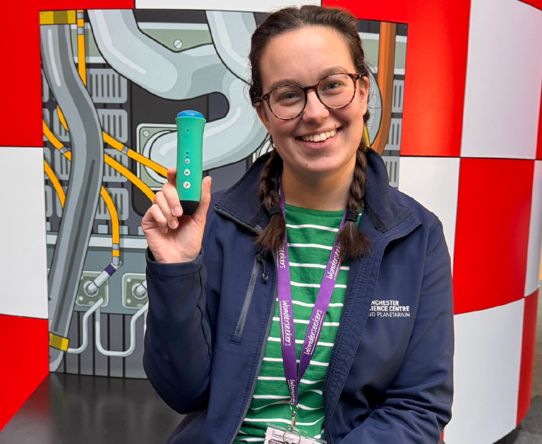 Jenny is sat on the large rocket exhibit, smiling. She has long dark brown hair that's been platted and is wearing her navy-blue Winchester Science Centre jacket. In her left hand is a Discovery Pen.