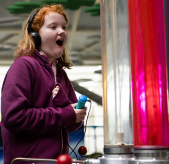 Girl with mid-length ginger hair is playing on  the Velocity exhibit. She is holding the Discovery Pen in her right hand and wearing a pair of black headphones on her head. She has an excited expression.