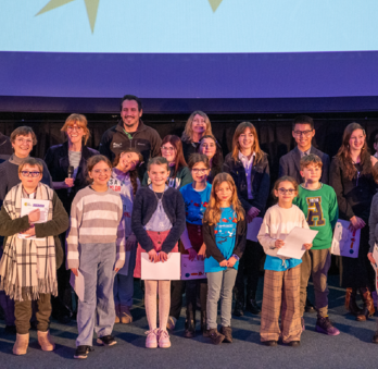 A group pf people are stood on a stage in front of a Planetarium screen. Adults are at the back and children are at the front. Some of the children are holding certificates.