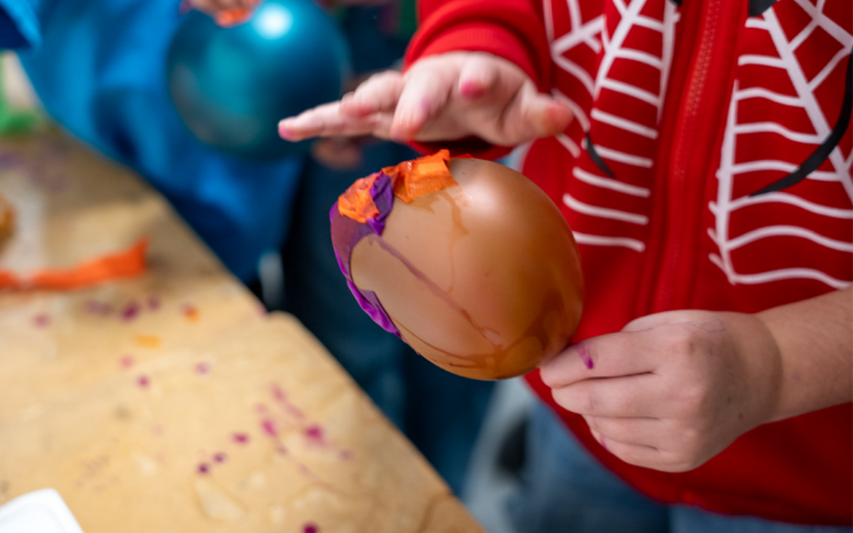 A child's hands can be seen holding a copper-coloured balloon in their left hand. With their right hand, they are covering the balloon with orange and purple tissue paper.