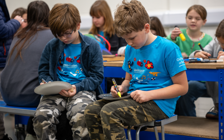 Two boys with short brown hair, wearing bright blue t-shirts camouflage trousers, are drawing constellations on black paper plates. They are both holding the plates on their laps.