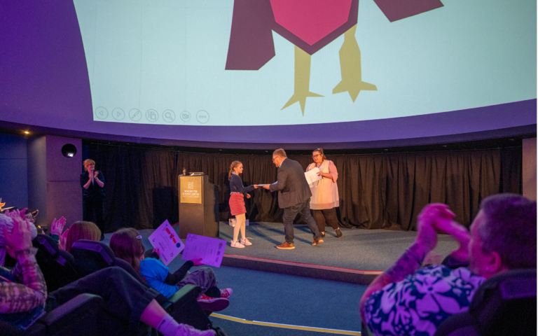 In the Planetarium, a girl with dark blonde platted hair is stood on the stage being handed a certificate by a man by a man with short hair, glasses, wearing a black suit. Everyone watching is clapping.