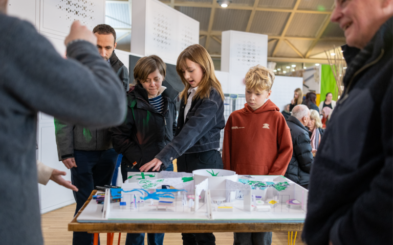 A family is stood looking at a carboard model of an exhibit on a table. A girl with mid-length light brown hair, wearing a white shirt and denim jacket, is pointing to the left of the model.