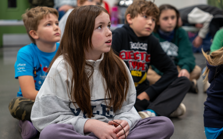 Children are sat cross-legged on the floor. At the front, is a girl with long dark brown hair, wearing a light grey jumper. She is sat with her hands in her lap and is looking slightly upwards with a look of concentration on her face. Behind her left shou