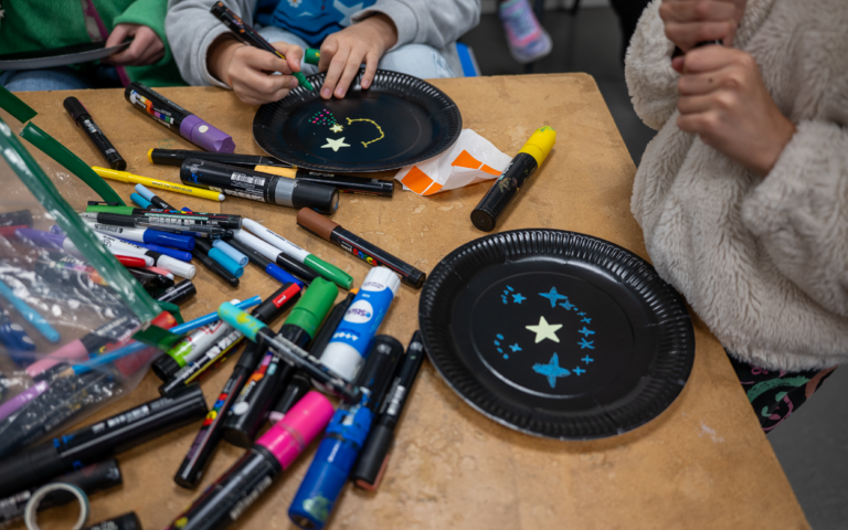 A wooden table with a selection of coloured markers and pens in a pile. There's two black paper plates, one with a yellow star sticker in the middle and bright blue star drawings around the edge. The other has two yellow star stickers in the middle with a