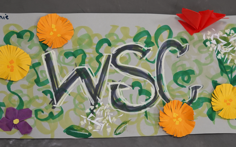 'WSC' is written in the centre of the page with green vines drawn around the edge and flowers stuck on the corners of the page.