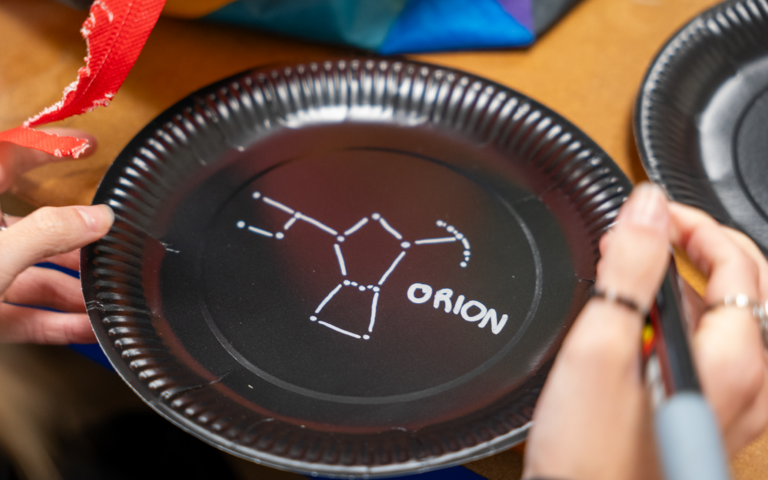 A black paper plate with a drawing of a constellation in white marker pen. To the right of the drawing is white writing which reads: "Orion".