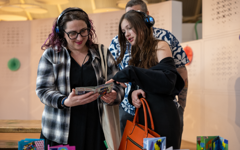 A girl with long dark brown hair, wearing a black top is showing her parents her artwork. The mother, who is wearing a black top, a black and white plaid jacket, and black glasses is stood next to the girl smiling down at the leaflet she’s holding.