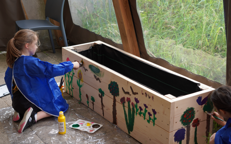 Two girls in blue aprons, painting nature related objects onto a large wooden planter