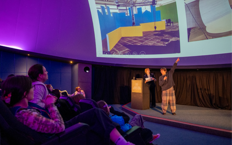 A women with short brown hair, wearing patchwork trousers and a dark grey jumper is stood on a stage. She is pointing to photos on the Planetarium screen. Stood to the right of her is a lady with ginger hair, holding a piece of white paper. People sat in 