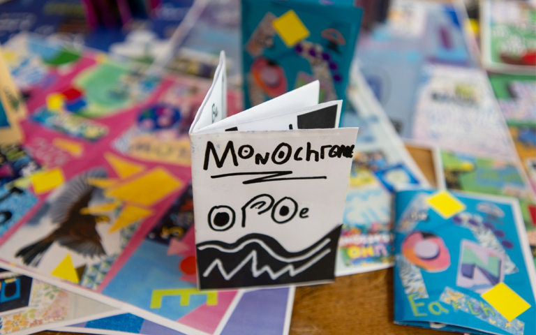 There’s a selection of colourful artwork spread across a table. In the centre of the table is a leaflet decorated with black and white patterns. On the front page is text that reads “monochrome” and “OPOE”.