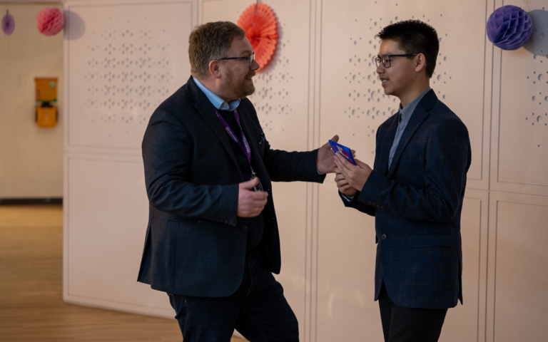 A boy with short black hair, wearing a black suit and black glasses is talking to the CEO of Wonderseekers. A man with short light brown hair, wearing a black suit with a purple Wonderseekers lanyard around his neck. They are both smiling enthusiastically