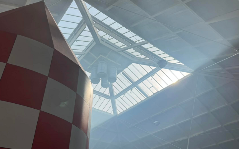 The roof of the pyramid building is filled with smoke. Sun is shinning through the windows and smoke. In the bottom left-hand corner is the top of the giant rocket exhibit.