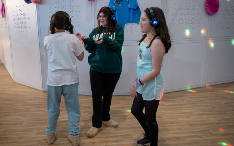 3 girls with long brown hair are stood talking in a group. They are all wearing blue headphones.