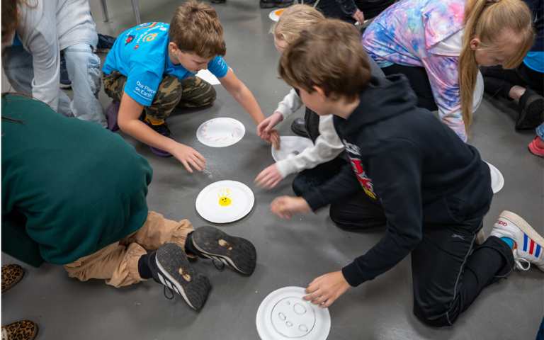 A group of children are knelt down on the floor reaching for white paper plates. The plates have the sun drawn on them.