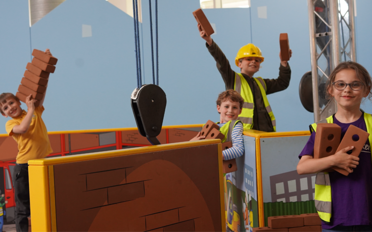 Children wearing high-vis jackets and yellow hard hats are smiling and playing on the crane exhibit